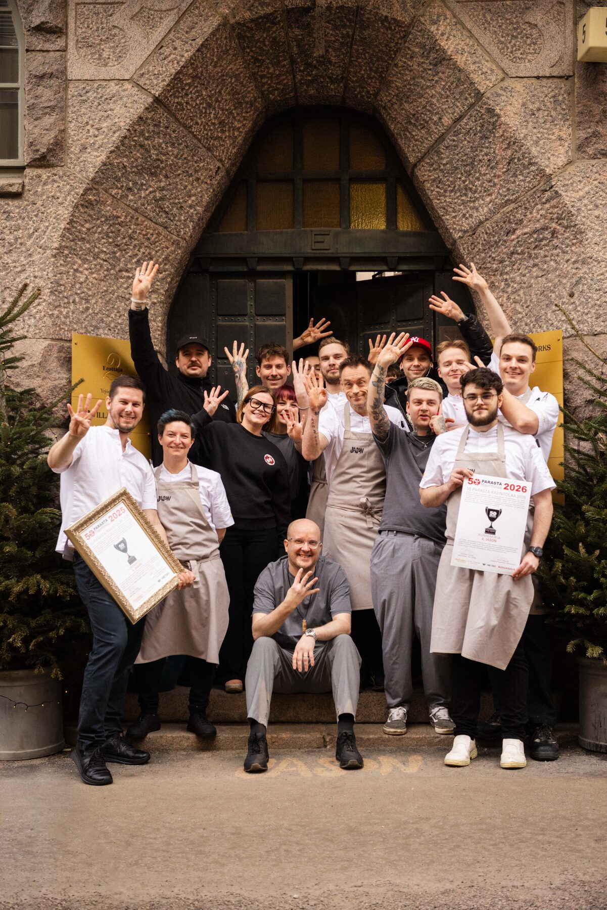 Jason restaurant team in front of the granite arched entrance, raising four fingers, holding 50 Parasta 2025 and 2026 award certificates.