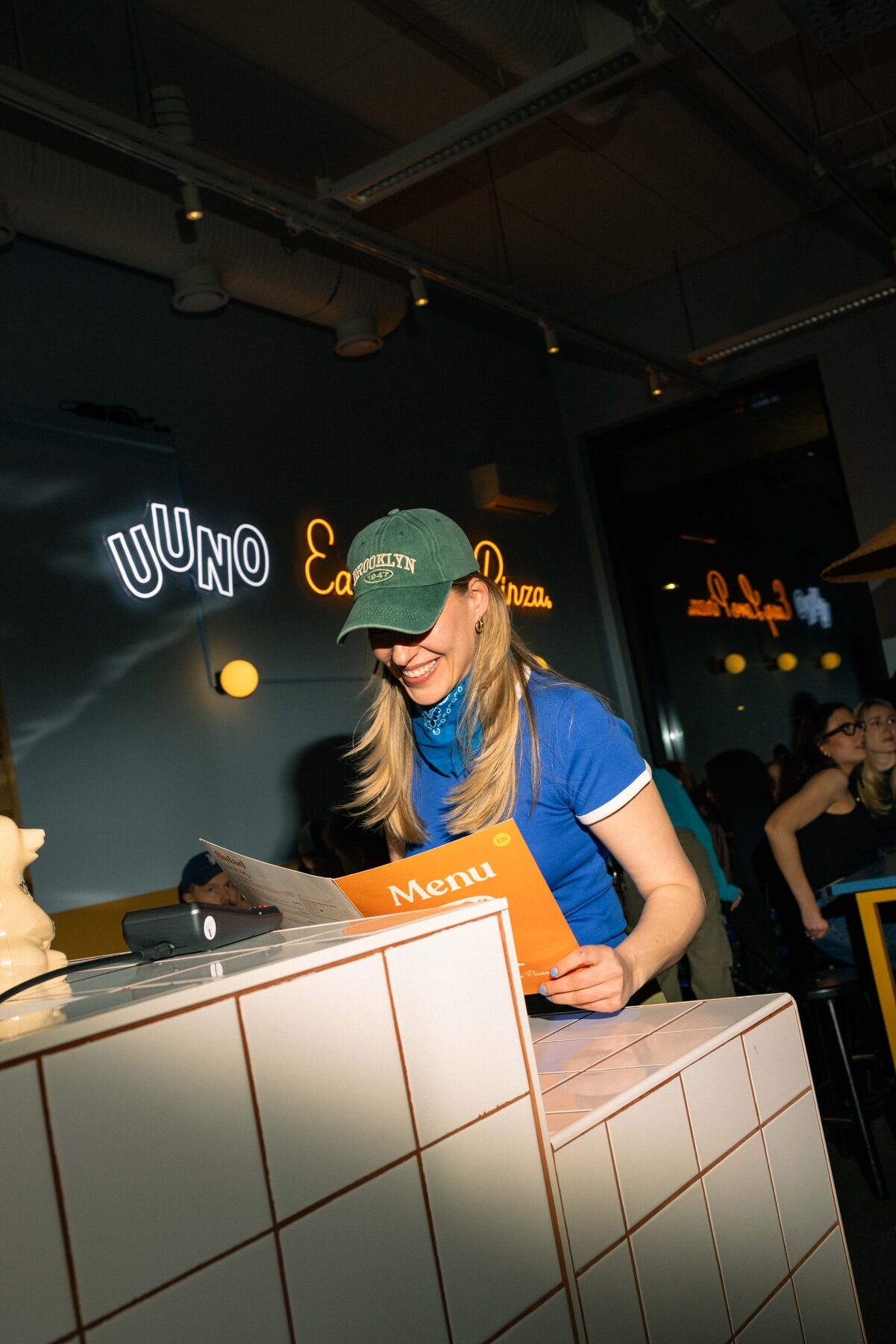 Uuno staff member in a green Brooklyn cap and blue retro shirt, holding an orange menu, neon UUNO sign in the background.